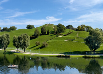 People enjoying Munich's Olympic Park grounds where there is a beautiful lake where boats can be rented which is surrounded by a picturesque grassy hill with a sweeping view.
