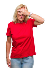 Middle age blonde woman over isolated background peeking in shock covering face and eyes with hand, looking through fingers with embarrassed expression.