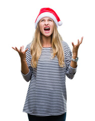 Young beautiful blonde woman wearing christmas hat over isolated background crazy and mad shouting and yelling with aggressive expression and arms raised. Frustration concept.