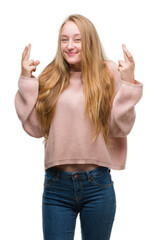 Blonde teenager woman wearing pink sweater smiling crossing fingers with hope and eyes closed. Luck and superstitious concept.