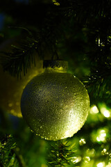 Glittery golden ball on artificial Christmas tree. Vertical greeting card template. Shiny bauble hanging on a branch of plastic Xmas tree in the dark, lit by garlands. Close-up, blurred background.