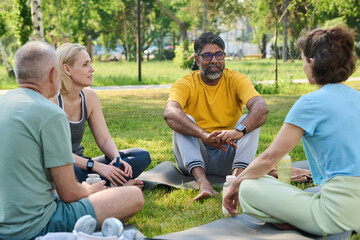 Naklejka premium Smiling mature man in activewear sitting on mat between diverse people and listening to elderly female yoga trainer at break