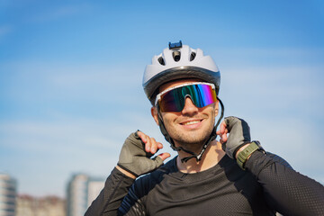 A cyclist in sports gear smiles while adjusting his helmet, wearing reflective sunglasses on a sunny day.