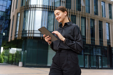 A young woman in a stylish black outfit uses a tablet while walking outside a modern glass building, smiling and focused.