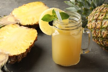 Tasty pineapple juice in mason jar, mint and fruits on grey textured table, closeup