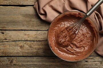 Chocolate dough in bowl and whisk on wooden table, top view. Space for text