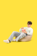 Young man in 3d eyeglasses sitting on pouf with popcorn on yellow background