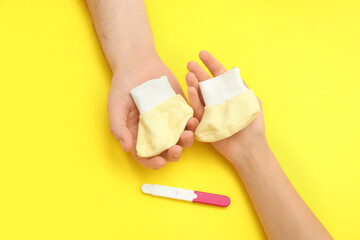 Hands of loving young couple with pregnancy test and baby booties on yellow background
