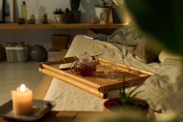 Bamboo tray with empty glass and teapot with hot herbal tea standing on edge of comfortable bed or couch with cushions