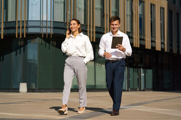Fototapeta premium A man and woman in business attire walk confidently outdoors, with the woman on a phone call and the man holding a tablet.