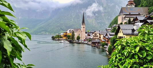 A picturesque view of Hallstatt, Austria, nestled by the tranquil lake and surrounded by towering misty mountains. The charming village is adorned with colorful houses and a prominent church, making i