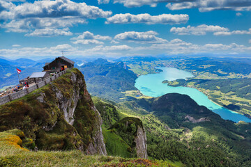 A breathtaking view from the Himmelspforte on the Schafberg mountain peak in Austria, overlooking a vibrant turquoise lake and a vast green valley. The landscape features a traditional alpine hut, cle