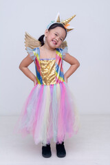 Full body photo of a joyful little asian girl dressed in a colorful unicorn costume with a golden horn and wings, smiling playfully. Isolated on a white background.