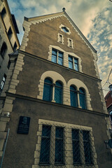 A close-up of a historic building in Riga, Latvia, showcasing its intricate brickwork and architectural details. The building's facade features a prominent arched window and a decorative plaque.