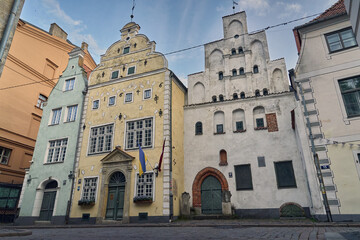 Fototapeta premium A picturesque view of the Three Brothers, a trio of historic buildings in Riga, Latvia. The cobblestone street, adorned with charming flowerpots, adds to the charming atmosphere. The buildings' intric