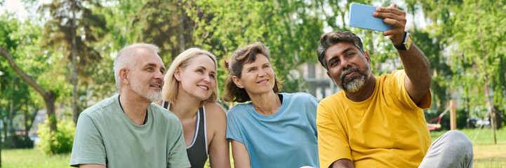 Obraz premium Banner of happy mature people in activewear looking at screen of mobile phone held by multiethnic man while watching online video outdoors