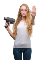 Blonde woman teenager filming holding super 8 video camera with open hand doing stop sign with serious and confident expression, defense gesture