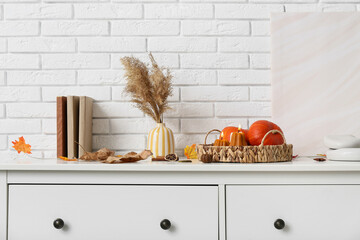 Wicker basket with burning candles in shape of pumpkins, autumn leaves and books on commode at home