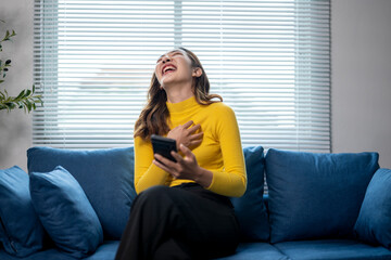 Obraz premium Happy young woman is sitting on sofa holding smartphone and laughing out loud