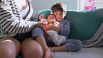 Mother hands newborn sibling to older brother seated on couch, highlighting the joy and tenderness of family moments and the arrival of a new family member