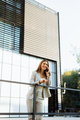 Fototapeta premium Smiling business woman enjoying a sunny day outdoors while texting on her smartphone near modern architecture in an urban setting