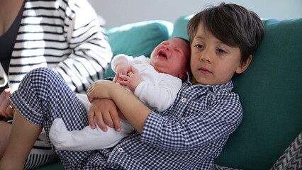 Young boy holding crying newborn sibling on the couch, emphasizing the protective and nurturing bond between siblings in a serene home setting