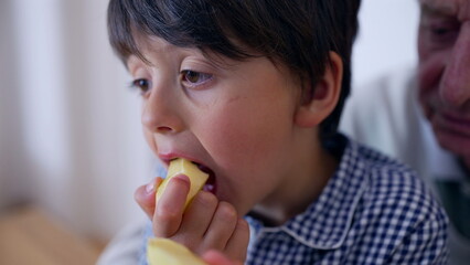 Young boy enjoying apple slice, close-up of eating, healthy snack, thoughtful expression, child eating, focus on food, home setting, family moment, concentrated child