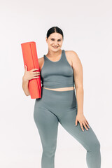 A confident young plump woman holds an unforgettable yoga mat in her hands on a light background, preparing for a workout. Ideal for fitness promotion, active lifestyle and wellness programs.