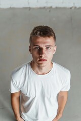 A young man with a shaved head and a white shirt is standing in front of a wall. He has a serious expression on his face