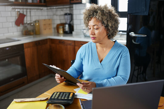 A concerned mature woman with curly gray hair reviews paperwork at her kitchen table. She holds a document while looking at it intently, surrounded by a laptop, calculator, and papers