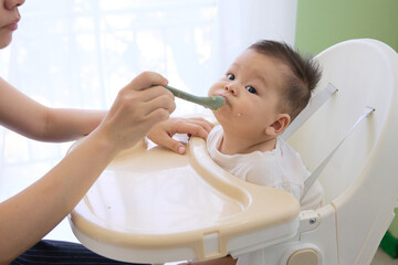 The baby sat in the dining chair with his mouth wide open, eating the soup his mother fed him