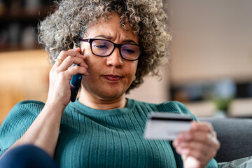 A worried woman holds her credit card and speaks on the phone, looking stressed. Her face is...