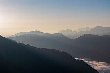 Serene alpine landscape at dawn in Gailtal Alps, Carinthia, Austria, Europe. Silhouette of majestic hazy mountain ridges against morning sky. Valley covered in fog, clouds. Peaceful serene atmosphere