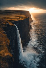 A dramatic waterfall cascades down a sheer cliff face into the churning sea as the sun sets over the horizon, casting a golden glow over the landscape.