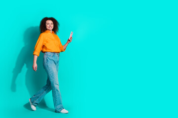 Naklejka premium Full size photo of positive optimistic woman dressed orange shirt hold smartphone go empty space isolated on teal color background