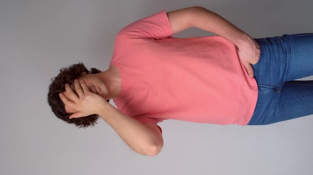 Ashamed young brunet curly man put hand on face facepalm epic fail mistaken omg gesture on gray background. Sincere emotions, emotional embarrassing confuse reaction. Guy wearing pink t-shirt, jeans.