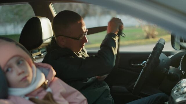 A man wearing a green jacket sits in the driver's seat, holding a phone and having just unbuckled his seat belt. a young girl in a pink knit hat and white scarf sits peacefully, looking out the window