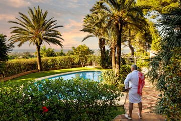 A man of retirement age in a white robe by the pool against an exotic landscape with palm trees and a pool enjoying the setting sun.