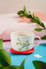 Ceramic cup with floral design. Tea bag tag on blue surface. Green leaves and twigs.