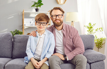 Portrait of smiling father and son wearing glasses sitting on sofa in cozy living room. Bearded man hugging his son with one hand posing for photo and looking into camera. Happy fatherhood concept. 