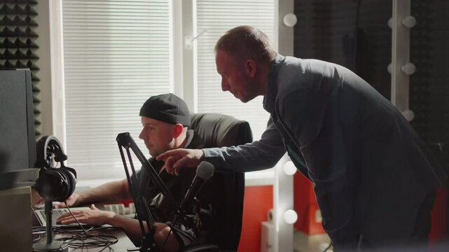 Two men are in a studio room, one seated in front of a computer, likely a producer, wearing black clothes, while the other, dressed in a long sleeve shirt, leans over, explaining something intently