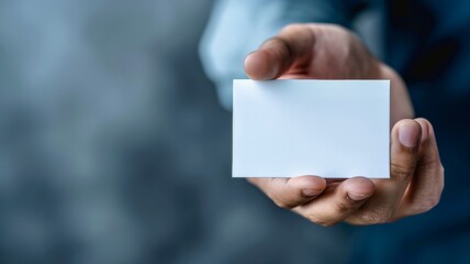 A person holding a blank business card in their left hand against a neutral textured background during daylight hours