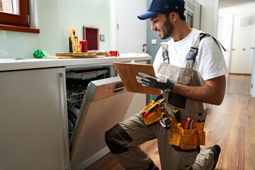 Service worker sitting near dishwasher and writing on clipboard in kitchen. High quality photo