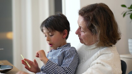 Grandmother attentively watching her grandson as he examines a piece of food, sitting on her lap. The boy is focused, showing a moment of curiosity and connection in a family setting