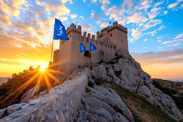 Fototapeta premium A medieval castle perched on a rocky hill, with banners flying and the sun setting behind it, casting a golden light over the scene