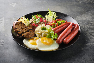 hearty breakfast featuring sunny side up eggs, grilled sausages, tomatoes, salads, and bread slices on a dark plate.