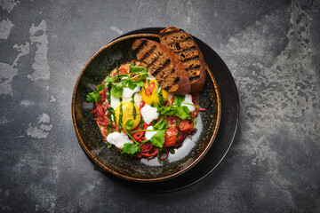 top view of a gourmet breakfast featuring eggs, grilled bread, and fresh vegetables, served in a dark bowl on a textured surface.
