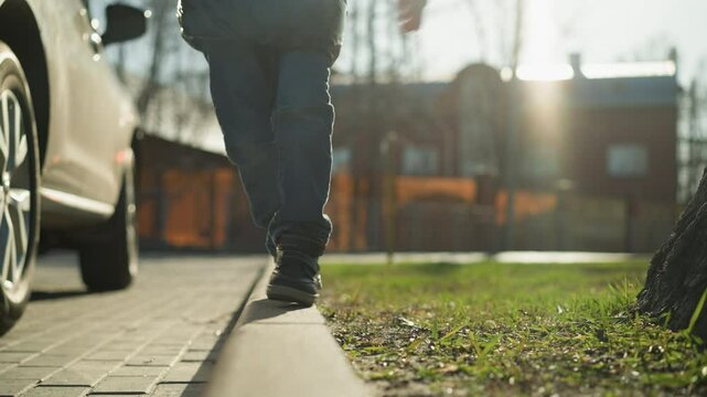 A close-up shot of a child walking along a pavement, holding a stick and missing a step while balancing, the child is dressed in a shiny black jacket