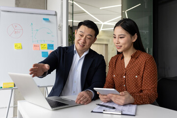 Asian business professionals engaged in collaborative work at office desk with laptop and tablet. Man explaining concepts using digital tools. Whiteboard with graphs and notes in background.
