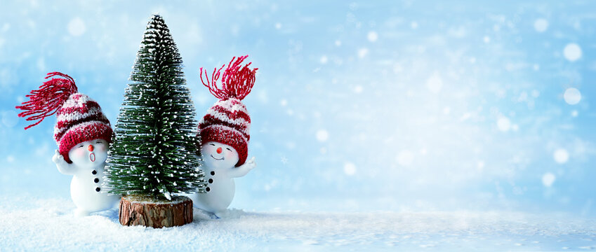 Two little snowmens in caps with Christmas tree on snow in the winter. Background with a funny snowman. Christmas card.